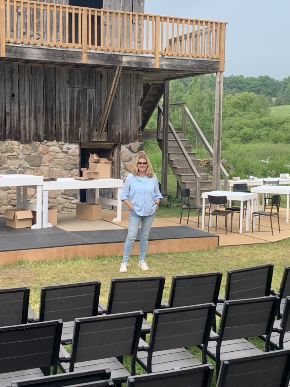 Cynthia stands on the outdoor stage looking into the empty seats, she wears sunglasses. 